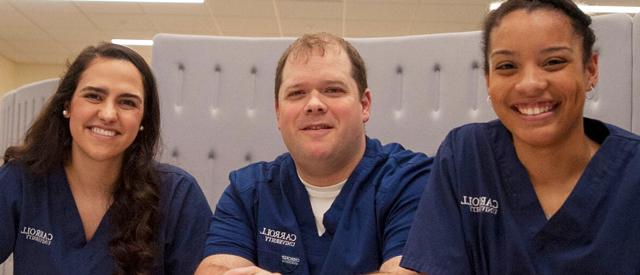 Three smiling healthcare professionals wearing scrubs with Carroll University's logo displayed.