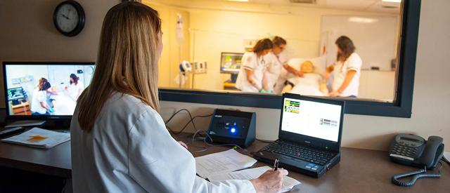 A clinical instructor observes nursing students practicing with a patient mannequin through a one-way mirror, while taking notes at her workstation.